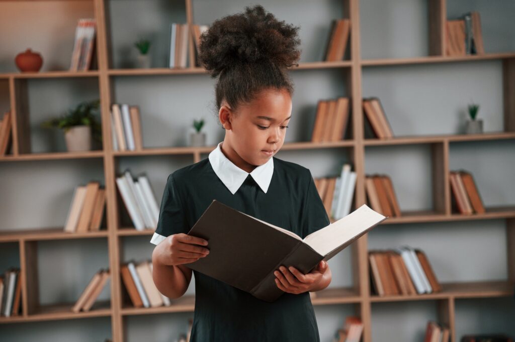 Holding book and reading. Cute african american girl in school uniform is at home library