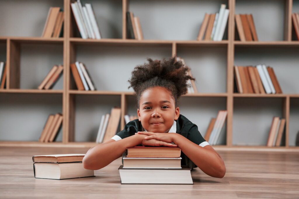 Sitting by the table with books. Cute african american girl in school uniform is at home library